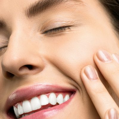 Gesicht einer lachenden Frau mit geschlossenen Augen, die ihre Fingerspitzen an ihre Wange hält